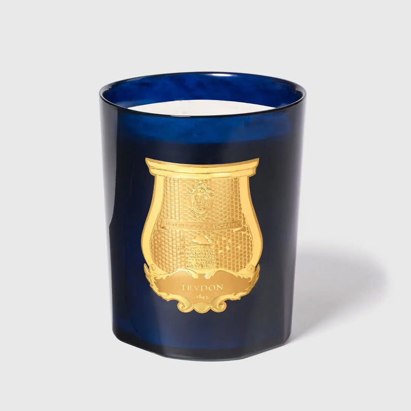 Vela aromática en vaso azul con emblema dorado en el frontal y mecha blanca
