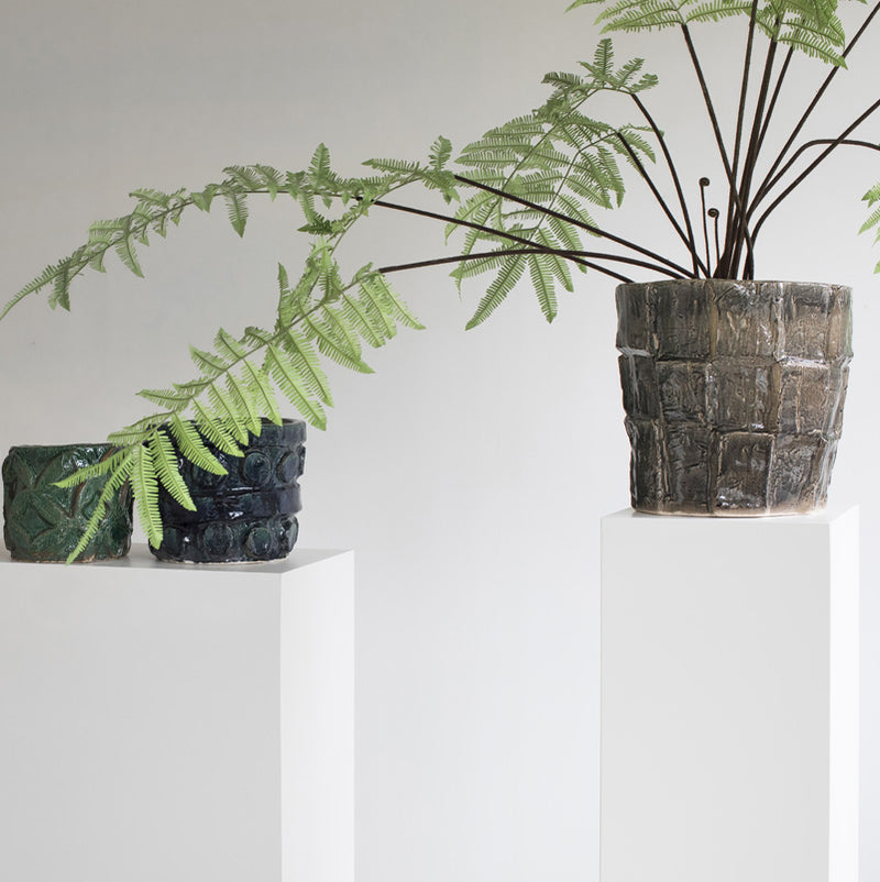 Macetero de cerámica gris oscuro con planta de hojas verdes sobre pedestal blanco