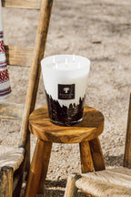 Vela blanca en vaso con diseño negro y llama encendida, sobre soporte de madera