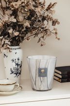 Vela en vaso de vidrio con vetas grises y etiqueta azul Baobab Collection, sobre mesa