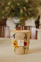 Vela en frasco de fibra tejida beige con flores amarillas y etiqueta frontal