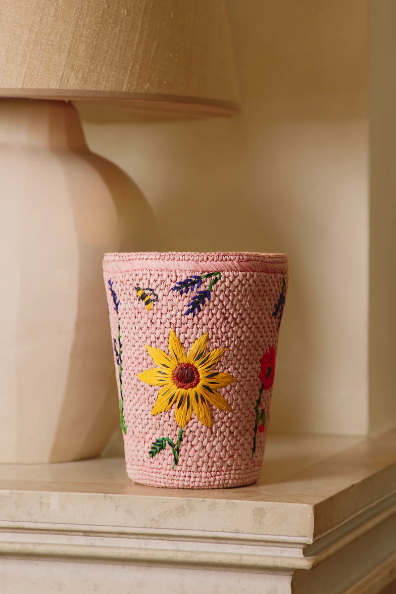 Vela Baobab en recipiente trenzado rosa con una flor bordada amarilla y etiqueta blanca.