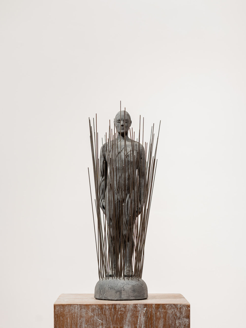 Escultura de busto con varillas metálicas verticales sobre base de piedra, gris oscuro