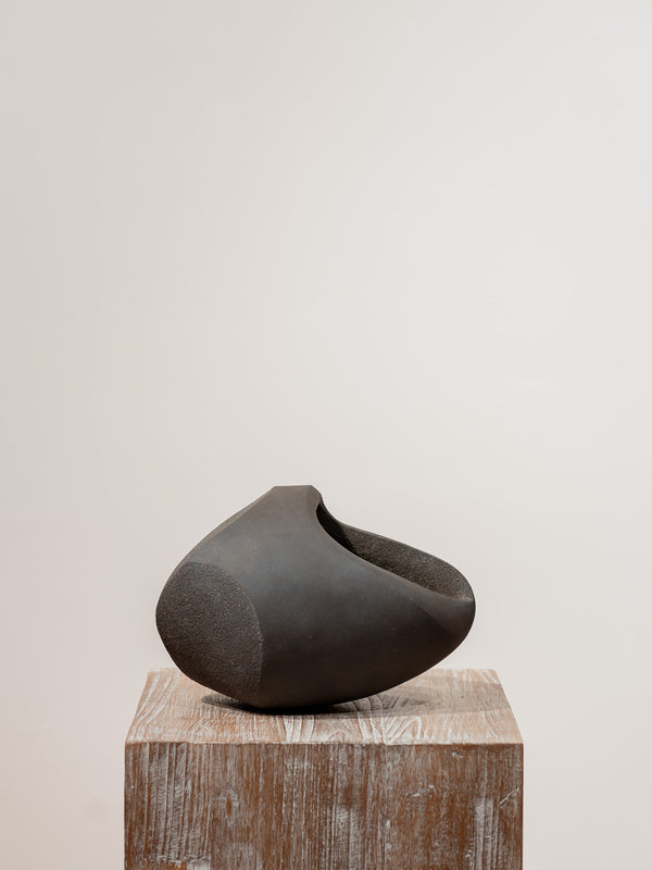 Escultura de cerámica gris oscuro con cavidad interior sobre base de madera