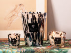 Trío de velas en vasos de vidrio con estampado negro y dorado; con mechas encendidas.